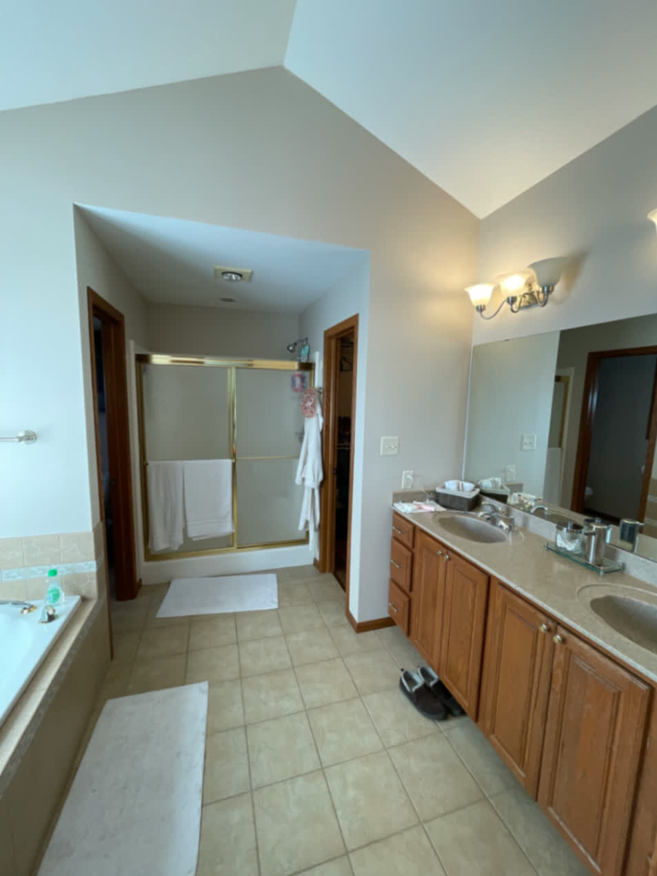 Bathroom before renovation in Cedar Falls with a built-in tub, gold-framed shower, double vanity, and beige tile floor