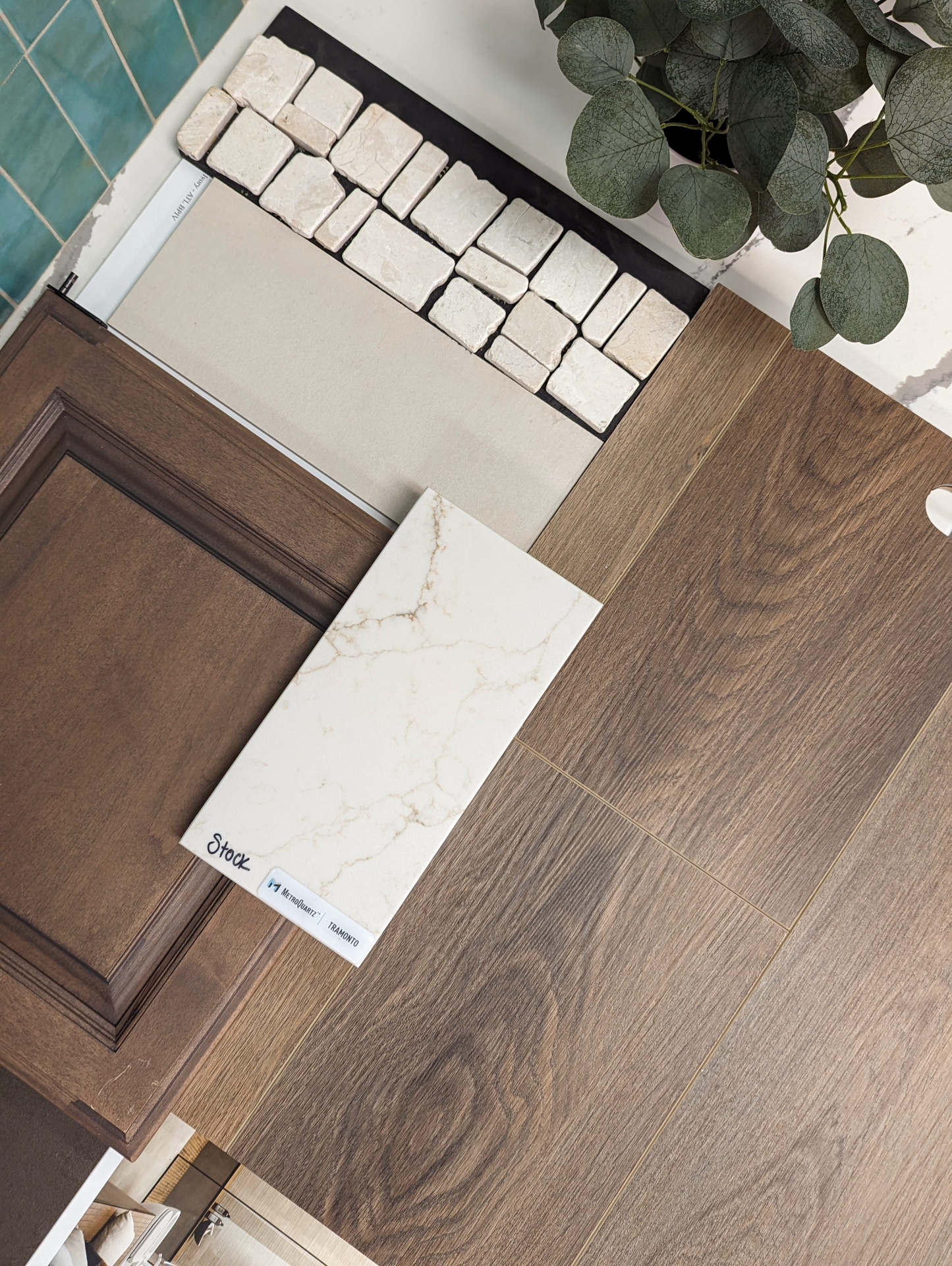 Material palette flat lay with wood-look flooring, quartz countertop sample, tumbled stone tile, teal accent tile, and wood cabinet door
