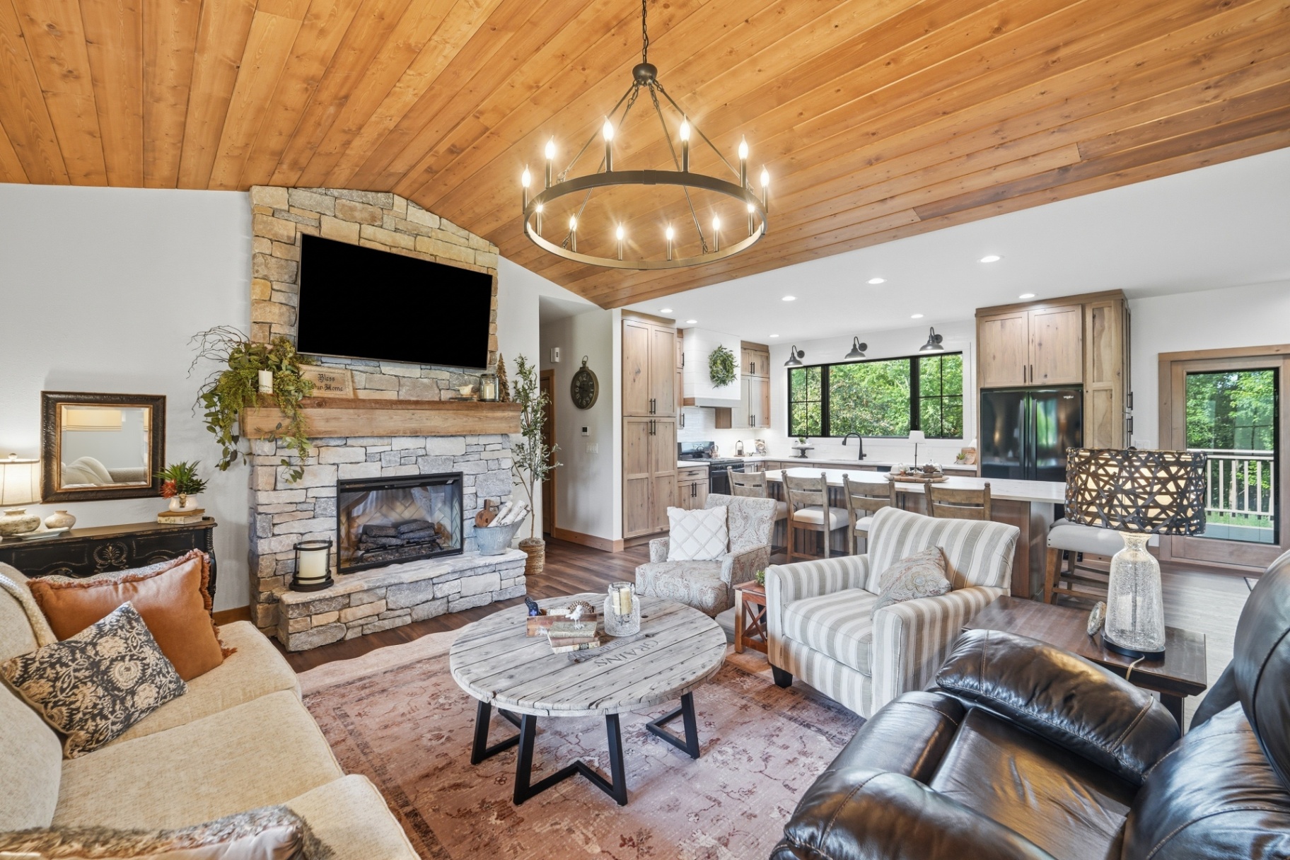 Open-concept living room with stone fireplace, wood ceiling, and view into the kitchen